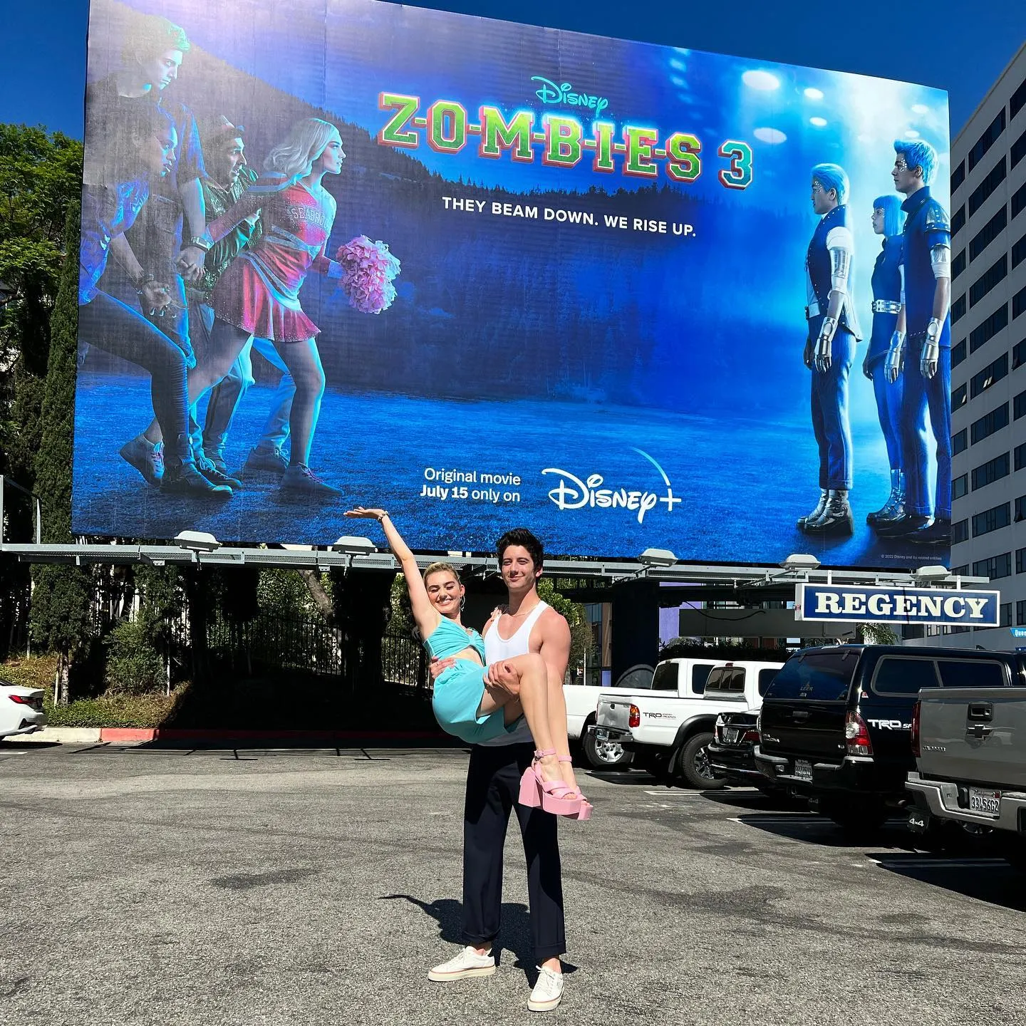 Milo Manheim and Meg Donnelly in front of Zombies 3's billboard poster